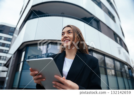 Young professional woman smiling while holding tablet outdoors in urban setting with modern architecture showcasing technology and lifestyle concept 132142659