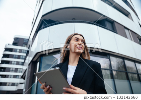 Confident businesswoman holding tablet while standing outside modern building with sleek architecture showcasing urban lifestyle concept Confident businesswoman holding tablet while standing outside modern building with sleek architecture showcasing urban lifestyle concept 132142660