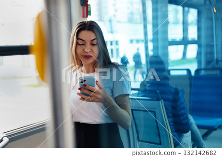 Young woman using smartphone in public transport setting, reflecting on glass, engaged in digital communication, modern lifestyle concept 132142682