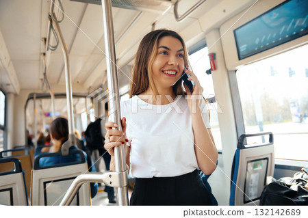 Young woman smiling while talking on smartphone in public transport setting with passengers around her showcasing urban lifestyle concept 132142689
