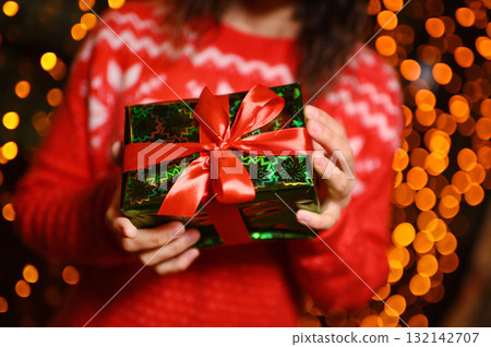 close-up - a girl in a red sweater with a Scandinavian pattern holds a Christmas present in her hands against the background of Christmas lights. close-up - a girl in a red sweater with a Scandinavian pattern holds a Christmas present in her hands against the background of Christmas lights. 132142707