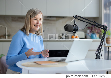 Senior woman smiling and gesturing while recording a podcast or livestream using a professional microphone and laptop in a bright home kitchen, communicating online Senior woman smiling and gesturing while recording a podcast or livestream using a professional microphone and laptop in a bright home kitchen, communicating online 132142838
