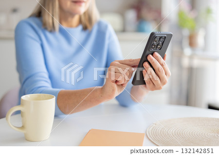 Senior woman is sitting at a white table, holding a black smartphone, touching the screen with her finger, engaging with digital content and technology in her modern home environment Senior woman is sitting at a white table, holding a black smartphone, touching the screen with her finger, engaging with digital content and technology in her modern home environment 132142851