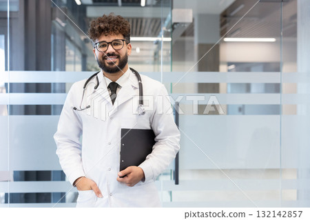 Young male doctor smiling, wearing a lab coat and stethoscope, holding a clipboard, standing in a contemporary clinic or hospital lobby, representing medical professionalism and healthcare Young male doctor smiling, wearing a lab coat and stethoscope, holding a clipboard, standing in a contemporary clinic or hospital lobby, representing medical professionalism and healthcare 132142857