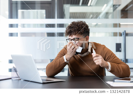 Young businessman suffering from a cold or allergies, blowing his nose with a tissue and holding a nasal spray while working at his desk in a modern office environment 132142878