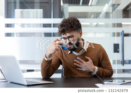 Young man experiencing an asthma attack and struggling with breathing difficulty at his workplace, using a blue inhaler while holding his chest with concern Young man experiencing an asthma attack and struggling with breathing difficulty at his workplace, using a blue inhaler while holding his chest with concern 132142879