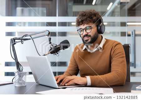 Young man wearing headphones and glasses sitting at a desk, typing on a laptop and speaking into a professional microphone, creating audio content for a podcast or online stream Young man wearing headphones and glasses sitting at a desk, typing on a laptop and speaking into a professional microphone, creating audio content for a podcast or online stream 132142885
