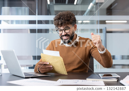 Smiling businessman excitedly opening an envelope with important news, celebrating success with a raised fist while sitting at his desk with a laptop and documents Smiling businessman excitedly opening an envelope with important news, celebrating success with a raised fist while sitting at his desk with a laptop and documents 132142887