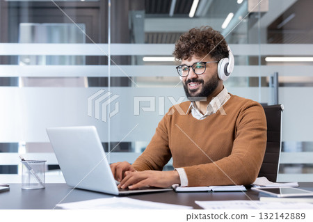 Smiling professional man with beard and glasses works on a laptop wearing white headphones at a modern corporate desk, focused and productive during a virtual meeting or training session Smiling professional man with beard and glasses works on a laptop wearing white headphones at a modern corporate desk, focused and productive during a virtual meeting or training session 132142889