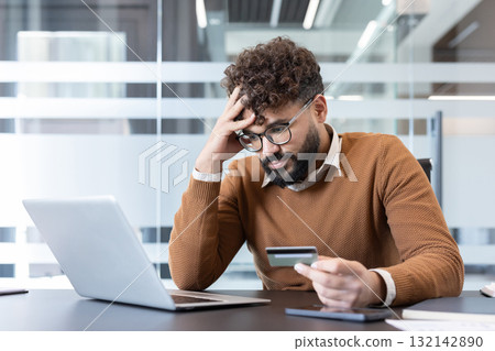 Young man feeling stressed and worried while looking at a credit card, experiencing financial difficulties and dealing with debt from online shopping on his laptop at the office 132142890