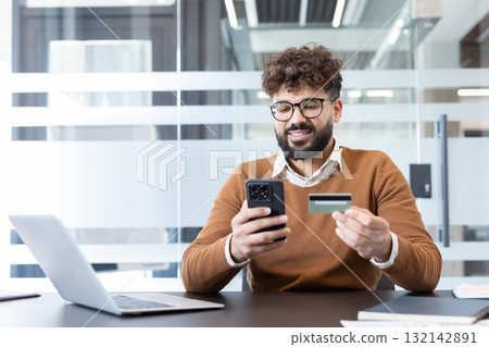Young man holding a smartphone and a credit card, confidently making an online payment or shopping transaction from his office desk, embracing modern e-commerce and digital finance Young man holding a smartphone and a credit card, confidently making an online payment or shopping transaction from his office desk, embracing modern e-commerce and digital finance 132142891