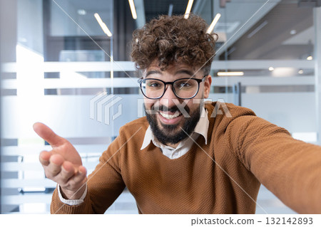 Happy young man with curly hair and beard enjoying a video call or online meeting in a contemporary office setting, actively communicating and gesturing towards the camera 132142893