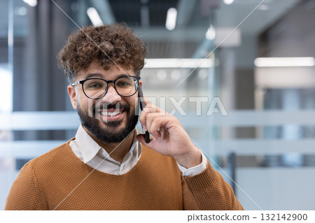 Smiling bearded man wearing glasses and a sweater, making a phone call on a smartphone while standing in a modern office with glass partitions, conveying successful communication Smiling bearded man wearing glasses and a sweater, making a phone call on a smartphone while standing in a modern office with glass partitions, conveying successful communication 132142900