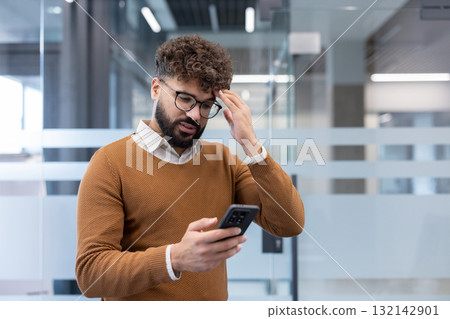 Man with a bearded face and eyeglasses holding a smartphone, experiencing headache and stress while reading bad news or difficult information in a modern office Man with a bearded face and eyeglasses holding a smartphone, experiencing headache and stress while reading bad news or difficult information in a modern office 132142901