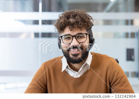 Male call center agent with a headset and glasses providing customer support, looking at the camera and smiling, representing communication, service, and professional assistance 132142904