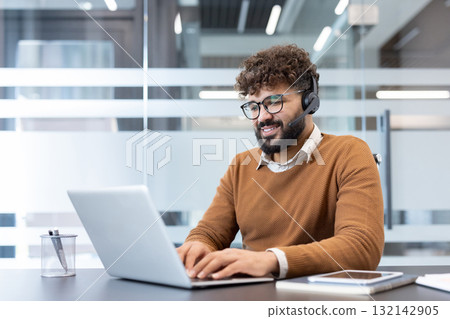 Smiling man wearing a headset and spectacles while typing on a laptop, providing customer support or engaging in online communication from a modern office environment Smiling man wearing a headset and spectacles while typing on a laptop, providing customer support or engaging in online communication from a modern office environment 132142905