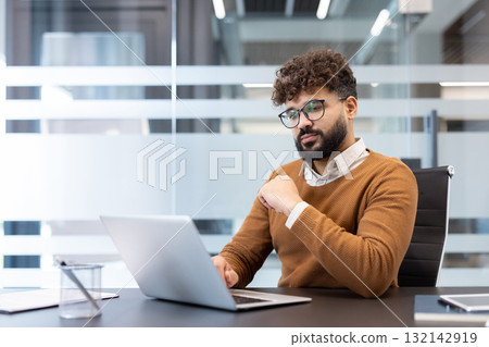 Young focused businessman working with a laptop at his desk in a blurred modern office environment, representing concentration, dedication, and professional productivity 132142919