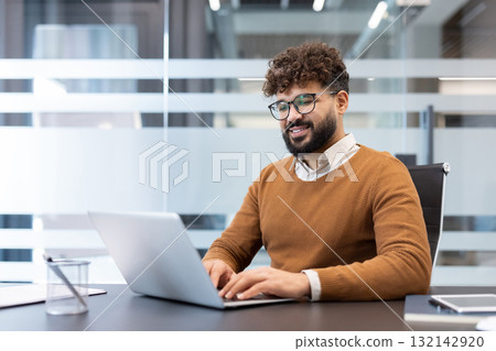 Young businessman typing on a laptop, smiling while confidently working at a desk in a contemporary corporate office, focusing on his professional tasks and digital workflow 132142920