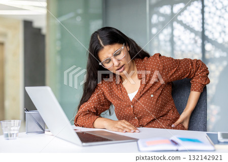 Young businesswoman experiencing back pain while sitting at her desk and working on a laptop, highlighting the physical strain of office work 132142921