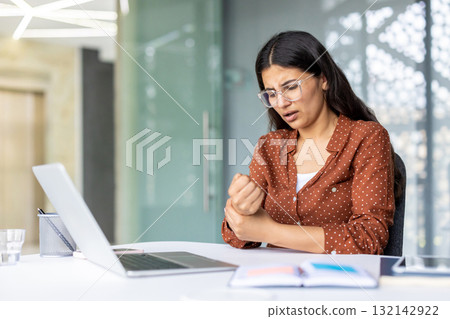 Young businesswoman experiencing wrist pain while working on a laptop in a modern office, highlighting the physical strain and health challenges associated with prolonged computer use 132142922