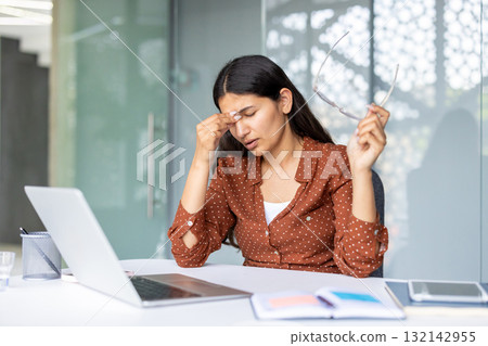 Stressed businesswoman taking off glasses and rubbing nose bridge while working on laptop in modern office, suffering from headache or eyestrain after long computer work 132142955