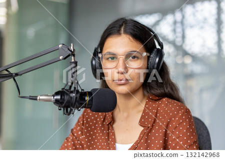 Young woman broadcaster wearing headphones and glasses, looking at the camera while sitting in a modern studio, recording a podcast or radio show using a professional microphone 132142968