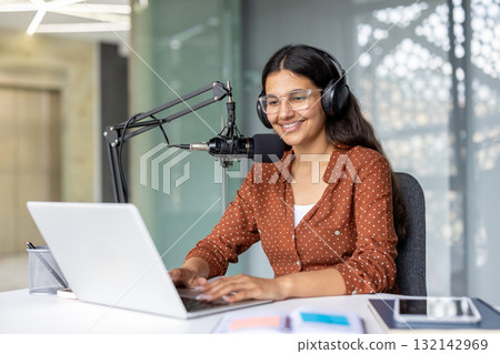 Young woman smiling with headphones, recording a podcast on laptop and pro microphone in a modern home office, confident content creator streaming and creating audio media 132142969