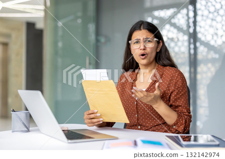 Young professional woman in glasses sits at her desk holding an envelope, reacting with shock and worry as she reads a distressing office letter or bad news notice Young professional woman in glasses sits at her desk holding an envelope, reacting with shock and worry as she reads a distressing office letter or bad news notice 132142974