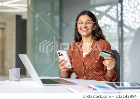 Happy businesswoman holding a credit card and using a smartphone, sitting at her desk in a modern office, enjoying online shopping and secure payment 132142984