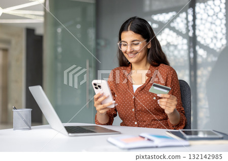 Young smiling woman sitting at a desk, enthusiastically shopping online, holding a credit card and smartphone, performing mobile payments or e-commerce transactions in a modern office Young smiling woman sitting at a desk, enthusiastically shopping online, holding a credit card and smartphone, performing mobile payments or e-commerce transactions in a modern office 132142985