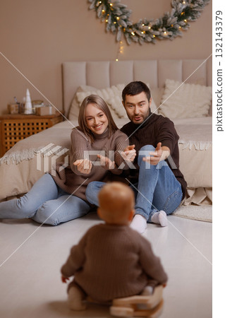 Young parents playing with baby on floor in cozy bedroom with beige wall and Christmas wreath 132143379