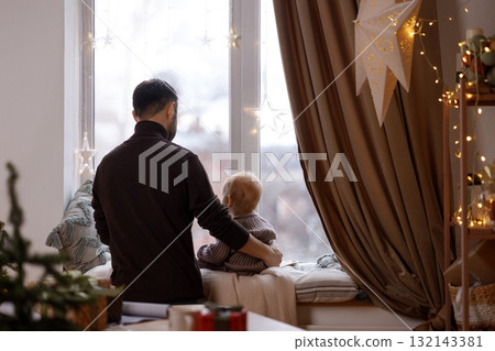 Father and baby looking out the window in decorated room with curtains and Christmas lights 132143381