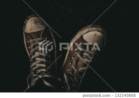 Worn brown sneakers with white laces against a dark background. A close-up shot of well-worn sneakers shows the details of the laces and the texture of the canvas. Dark key dramatic moody shot. 132143608
