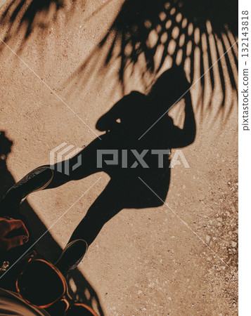 Silhouette of a person with palm tree shadows on a textured ground surface. A woman casts her shadow on a garden floor, contrast palm tree leaves adding depth and visual interest to the composition. Silhouette of a person with palm tree shadows on a textured ground surface. A woman casts her shadow on a garden floor, contrast palm tree leaves adding depth and visual interest to the composition. 132143818
