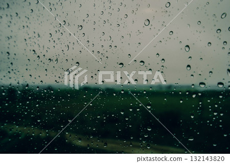 Raindrops on a wet glass window with blurred green landscape in the background. Moody rainy day, nature melancholy, peaceful atmosphere, seasonal weather and emotional calmness. Autumn fall season. 132143820
