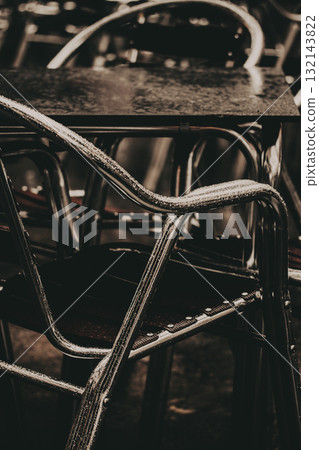 Close-up of wet metal chairs and a table in a moody, atmospheric restaurant setting. Cafe terrace after rain, creating a dark moody atmosphere Close-up of wet metal chairs and a table in a moody, atmospheric restaurant setting. Cafe terrace after rain, creating a dark moody atmosphere 132143822