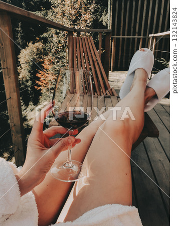 Relaxing moment on a wooden terrace with a glass of red wine in hand, legs in cozy slippers stretched out, enjoying calm nature views and warm sunlight in a peaceful outdoor setting. Travel girl. 132143824