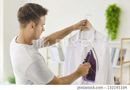 Man ironing white shirt with steam iron at home, smoothing wrinkles on hanger Man ironing white shirt with steam iron at home, smoothing wrinkles on hanger 132145104