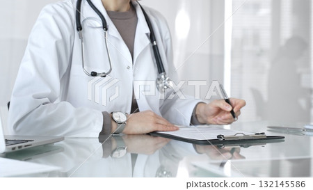 Female doctor in white coat with stethoscope writing patient notes on clipboard, completing medical records in clinic office. Medicine and health care 132145586