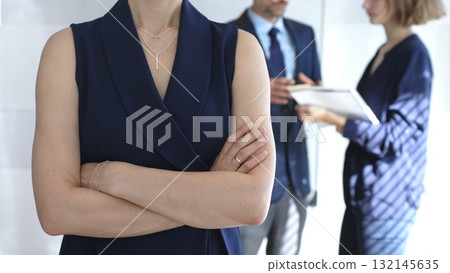 Professional businesswoman stands confidently with arms crossed in sharp focus as colleagues interact behind her during an office meeting, symbolizing leadership and corporate success Professional businesswoman stands confidently with arms crossed in sharp focus as colleagues interact behind her during an office meeting, symbolizing leadership and corporate success 132145635