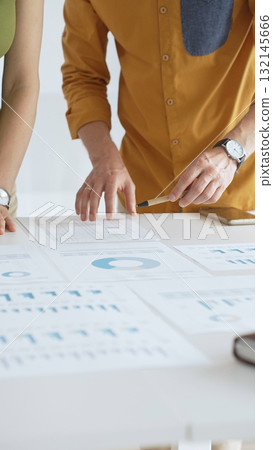 Business professionals analyzing financial reports, pointing at data points on charts and graphs lying across white desk surface 132145666