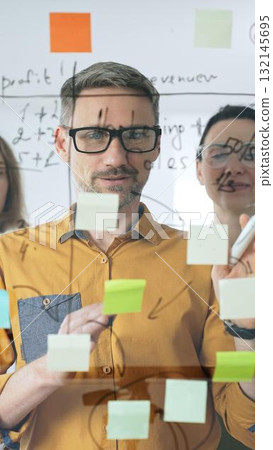 Professional team strategizing, writing collaborative notes on glass board, mapping innovative project workflow with marker Professional team strategizing, writing collaborative notes on glass board, mapping innovative project workflow with marker 132145695