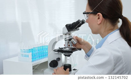 Female scientist examining microscope slide, analyzing chemical sample in sterile laboratory environment, performing precision medical research. Medicine and science concept 132145702