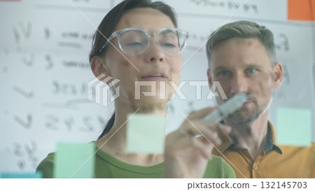 Professional businesswoman gesturing with marker, discussing strategic plans near glass board covered in colorful sticky notes during collaborative corporate meeting. Business people concept Professional businesswoman gesturing with marker, discussing strategic plans near glass board covered in colorful sticky notes during collaborative corporate meeting. Business people concept 132145703