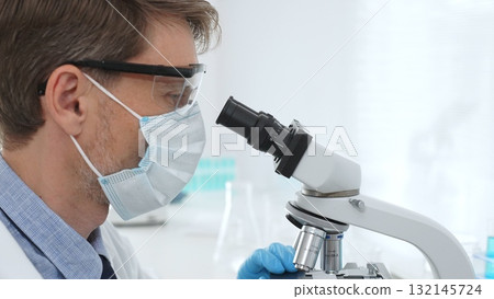 Scientist wearing mask and gloves using microscope doing scientific research in a laboratory with test tubes containing blue liquid. Medicine, healthcare and science concept Scientist wearing mask and gloves using microscope doing scientific research in a laboratory with test tubes containing blue liquid. Medicine, healthcare and science concept 132145724