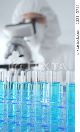 Close up of test tubes with blue liquid opposite a scientific researcher wearing protective gear tuning microscope lenses in laboratory. Medicine, healthcare and science concept Close up of test tubes with blue liquid opposite a scientific researcher wearing protective gear tuning microscope lenses in laboratory. Medicine, healthcare and science concept 132145732