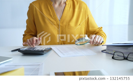 Female accountant wearing yellow shirt is examining a financial report with a magnifying glass while using a calculator and laptop in an office setting, close up. Audit and taxes in business Female accountant wearing yellow shirt is examining a financial report with a magnifying glass while using a calculator and laptop in an office setting, close up. Audit and taxes in business 132145755