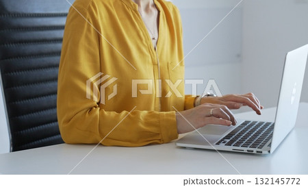 Businesswoman wearing yellow shirt sitting at white desk typing on laptop in bright modern office, working productively on computer. Business people concept Businesswoman wearing yellow shirt sitting at white desk typing on laptop in bright modern office, working productively on computer. Business people concept 132145772