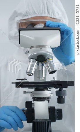 Male virologist in a hazmat suit, mask, and blue gloves, diligently working at a microscope in a laboratory researching viruses. Medicine, science concept 132145781