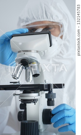 Male virologist in a hazmat suit, mask, and blue gloves, diligently working at a microscope in a laboratory researching viruses. Medicine, science concept 132145783